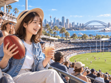 オーストラリア スポーツ観戦を楽しむ日本人留学生。晴れたスタジアムの客席で楕円形のボールを手に笑顔で応援している